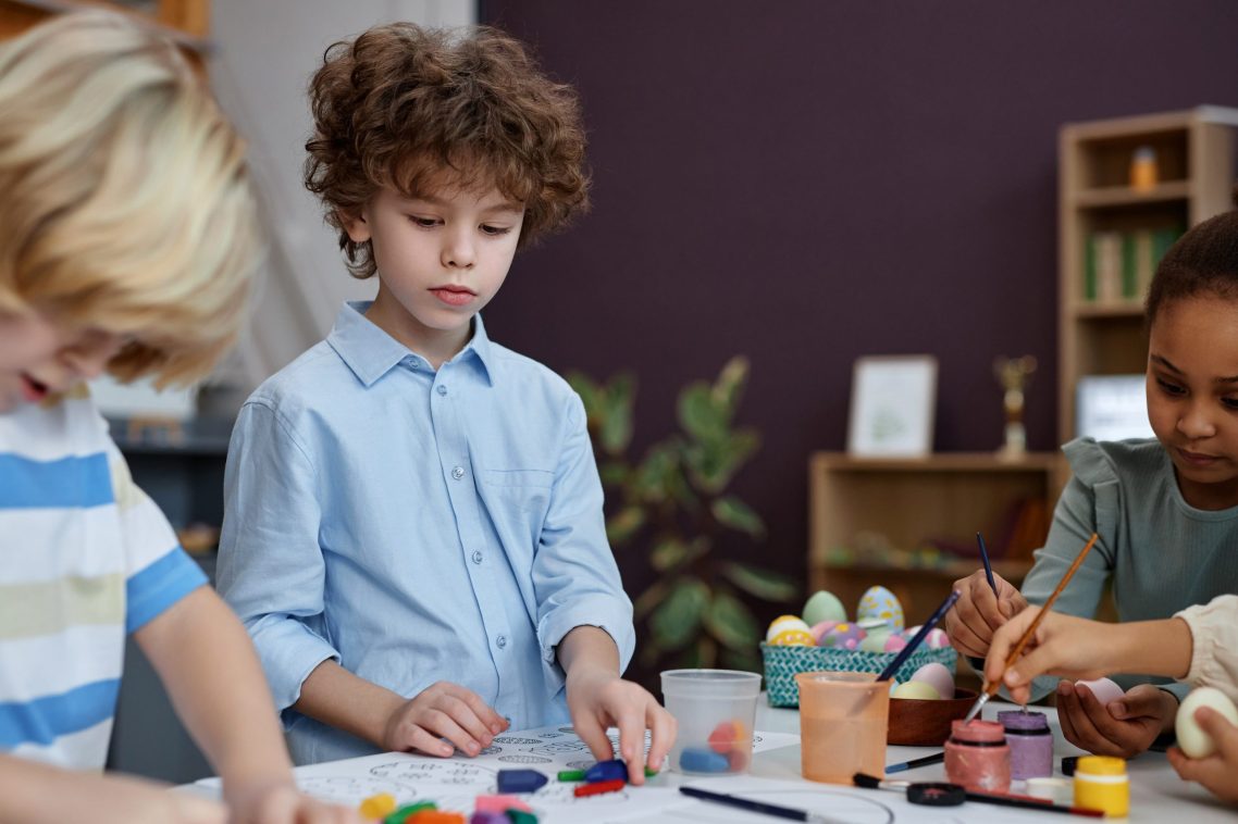 Portrait of cute curly haired boy coloring Easter pictures in preschool, copy space